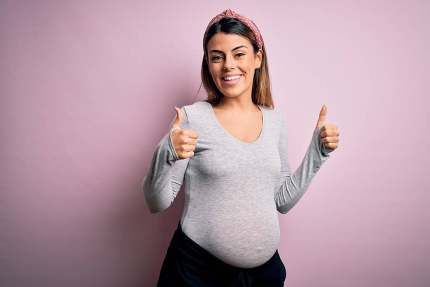 Jonge mooie brunette vrouw zwanger verwacht baby over geïsoleerde roze achtergrond succes teken doen positieve gebaar met de hand, duimen omhoog glimlachend en gelukkig. vrolijke uitdrukking en winnaar gebaar. - Foto, afbeelding