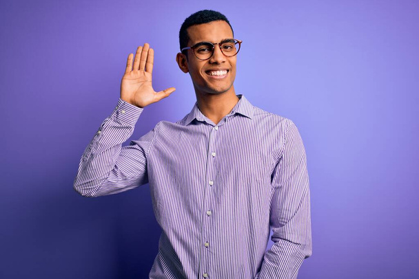 Bonito homem afro-americano vestindo camisa listrada e óculos sobre fundo roxo Waiving dizendo olá feliz e sorridente, gesto de boas-vindas amigável
 - Foto, Imagem