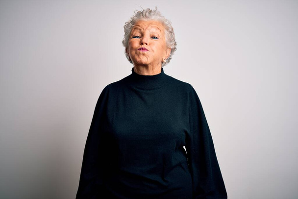 Senior beautiful woman wearing casual black sweater standing over isolated white background puffing cheeks with funny face. Mouth inflated with air, crazy expression. - 写真・画像