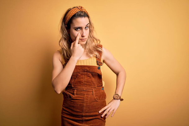 Young beautiful blonde woman wearing overalls and diadem standing over yellow background Pointing to the eye watching you gesture, suspicious expression - Photo, Image