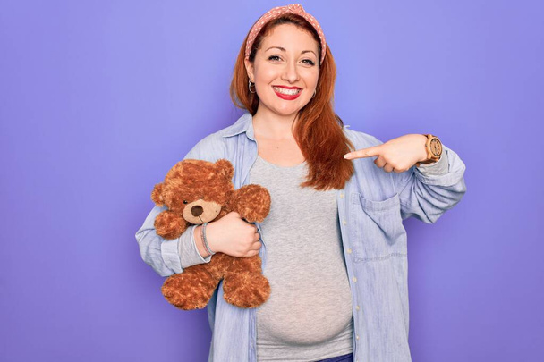 Young redhead pregnant woman expecting baby holding teddy bear over purple background with surprise face pointing finger to himself - Фото, изображение