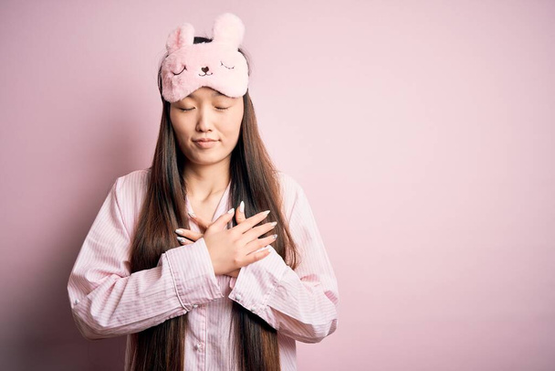 Young asian woman wearing pajama and sleep mask over pink isolated background smiling with hands on chest with closed eyes and grateful gesture on face. Health concept. - Fotó, kép
