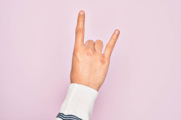 Hand of caucasian young man showing fingers over isolated pink background gesturing rock and roll symbol, showing obscene horns gesture - Foto, imagen