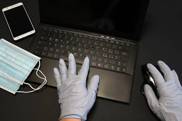 człowiek pracujący nad laptopem i noszący lateksowe rękawiczki do zapobiegania wirusom. Człowiek pracujący w domu za pomocą smartfona i laptopa. Niebieskie maski chirurgiczne na twarz dla koronawirusa, ochrona przeciw kovidowi-19. - Zdjęcie, obraz