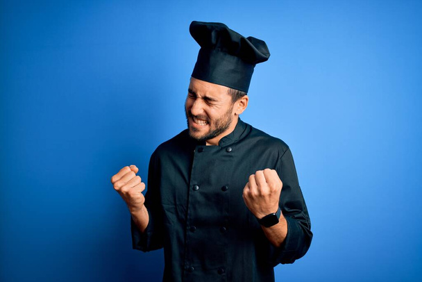 Jeune beau chef homme avec barbe portant uniforme de cuisinière et chapeau sur fond bleu très heureux et excité de faire geste gagnant avec les bras levés, souriant et criant pour le succès. Concept de célébration
. - Photo, image