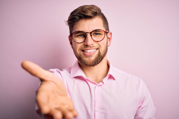Young handsome blond man with beard and blue eyes wearing pink shirt and glasses smiling friendly offering handshake as greeting and welcoming. Successful business. - Foto, immagini