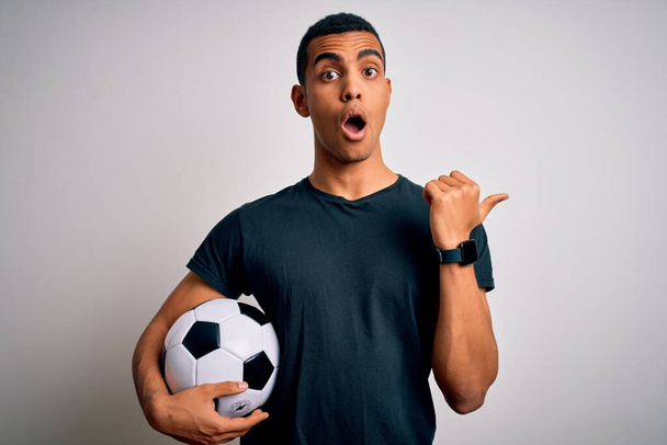 Fešák africký Američan hrající fotbal držící fotbalový míč na bílém pozadí Překvapený ukazující ukazováčkem na stranu, otevřená ústa užaslý výraz. - Fotografie, Obrázek