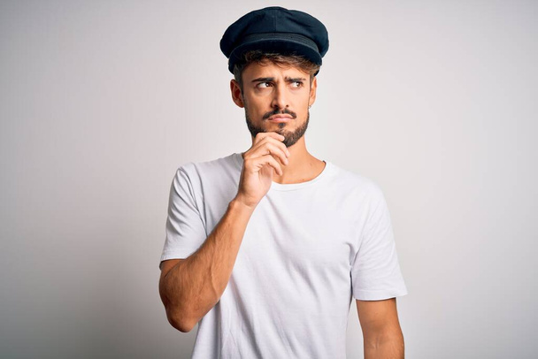 Young driver man with beard wearing hat standing over isolated white background with hand on chin thinking about question, pensive expression. Smiling with thoughtful face. Doubt concept. - Foto, Bild