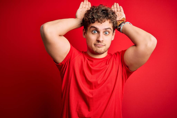 Young blond handsome man with curly hair wearing casual t-shirt over red background Doing bunny ears gesture with hands palms looking cynical and skeptical. Easter rabbit concept. - Photo, Image