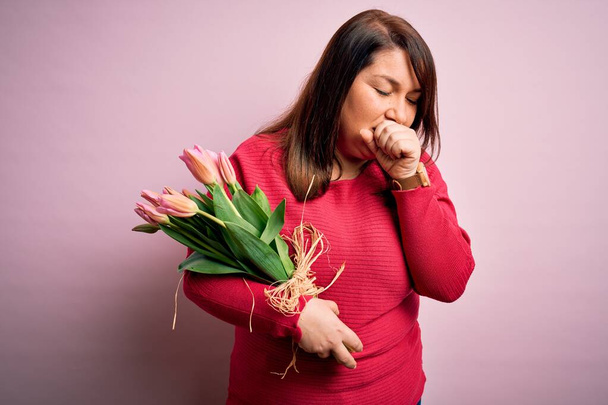 Belle femme de taille plus tenant bouquet romantique de fleurs de tulipes naturelles sur fond rose se sentant mal à l'aise et la toux comme symptôme de rhume ou de bronchite. Concept de soins de santé
. - Photo, image