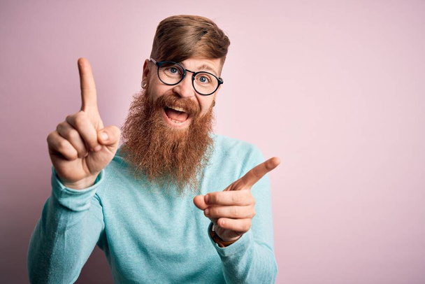 Красивый ирландский рыжий мужчина с бородой в очках на розовом изолированном фоне улыбается уверенно указывая пальцами в разные стороны. Копирование места для рекламы
 - Фото, изображение