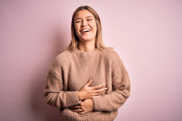Joven hermosa mujer rubia con suéter de lana de invierno sobre fondo aislado rosa sonriendo y riendo en voz alta porque broma loca divertida con las manos en el cuerpo
. - Foto, Imagen