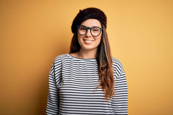 Young beautiful brunette woman wearing french beret and glasses over yellow background with a happy and cool smile on face. Lucky person. - Foto, afbeelding