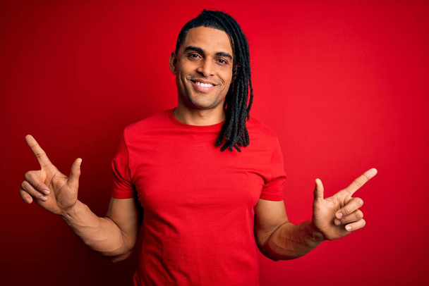 Ein junger gutaussehender afroamerikanischer Afro-Mann mit Dreadlocks, der ein rotes lässiges T-Shirt trägt und selbstbewusst lächelt und mit den Fingern in verschiedene Richtungen zeigt. Kopierfläche für Werbung - Foto, Bild