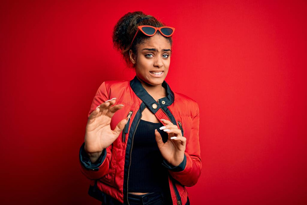 Beautiful african american girl wearing red jacket and sunglasses over isolated background disgusted expression, displeased and fearful doing disgust face because aversion reaction. With hands raised - Foto, Imagem