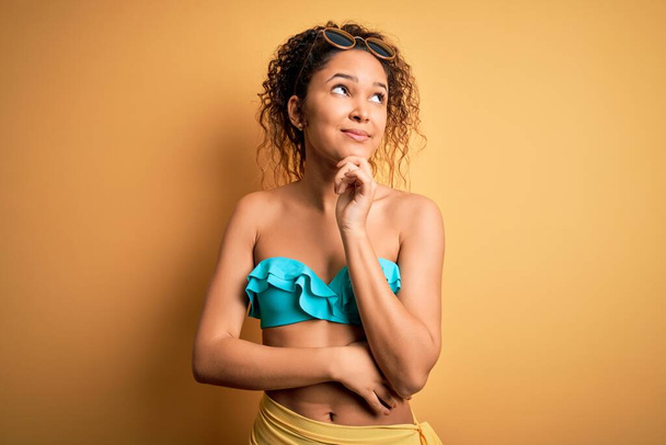 Young beautiful american woman on vacation wearing bikini over isolated yellow background with hand on chin thinking about question, pensive expression. Smiling and thoughtful face. Doubt concept. - Photo, Image