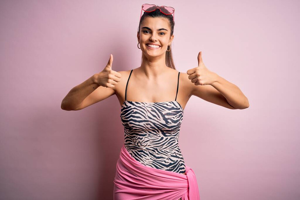 ピンクの背景に水着を着て休暇中の若い美しいブルネットの女性成功サインは手で積極的なジェスチャーをし、親指を上げて笑顔と幸せ。陽気な表情と勝者のジェスチャー. - 写真・画像