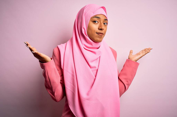 Mladá afro afro žena nosí muslimský hidžáb přes izolované růžové pozadí bezradný a zmatený výraz s rukama a zvednutýma rukama. Pochybný koncept. - Fotografie, Obrázek