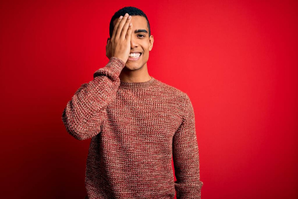 Young handsome african american man wearing casual sweater standing over red background covering one eye with hand, confident smile on face and surprise emotion. - Φωτογραφία, εικόνα