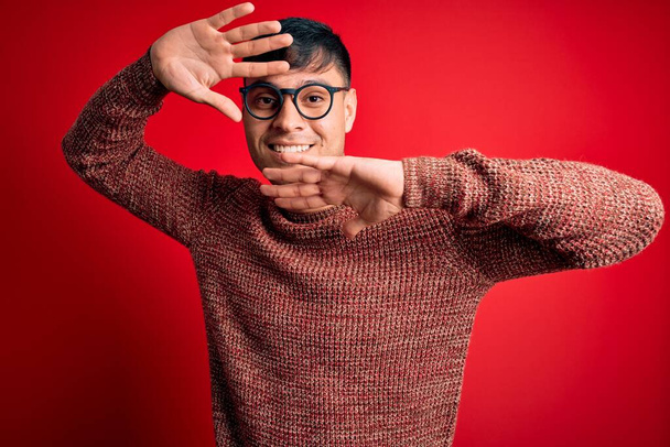 Young handsome hispanic man wearing nerd glasses over red background Smiling cheerful playing peek a boo with hands showing face. Surprised and exited - Foto, afbeelding