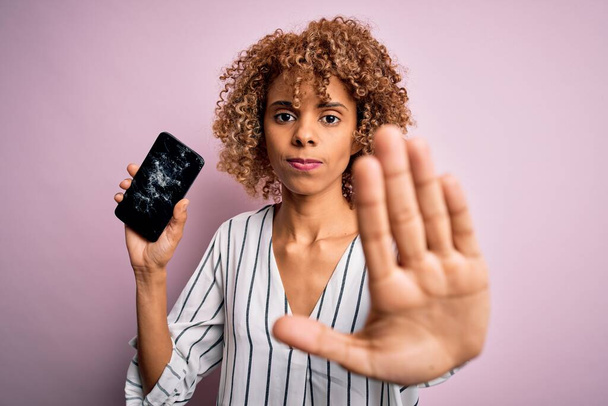 Jeune afro-américaine bouclée femme tenant smartphone cassé montrant écran fissuré avec la main ouverte faisant stop signe avec expression sérieuse et confiante, geste de défense
 - Photo, image