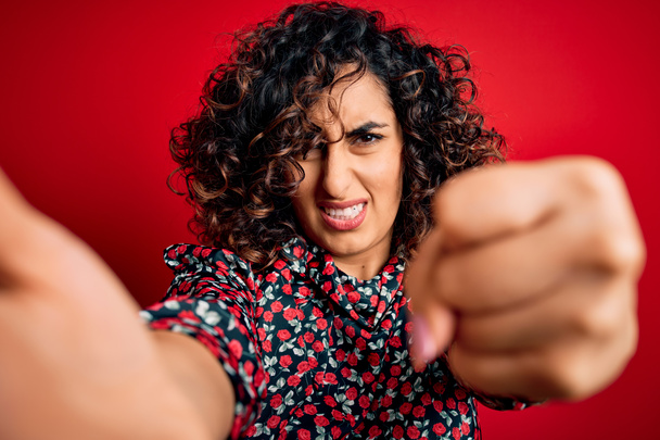 Young beautiful curly arab woman wearing dress making selfie by camera over red background annoyed and frustrated shouting with anger, crazy and yelling with raised hand, anger concept - Photo, Image