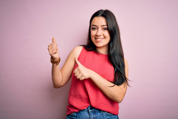 Mladá brunetka žena na sobě ležérní letní košile přes růžové izolované pozadí ukazuje na zadní straně s rukou a palci nahoru, s úsměvem sebevědomě - Fotografie, Obrázek
