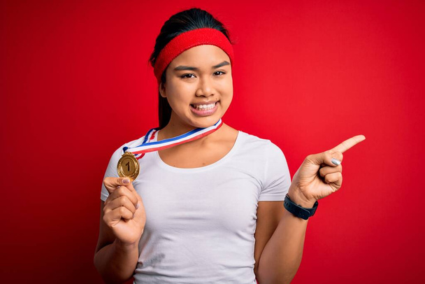 Joven asiática campeona chica ganando medalla de pie sobre aislado rojo fondo muy feliz señalando con la mano y el dedo a un lado
 - Foto, Imagen