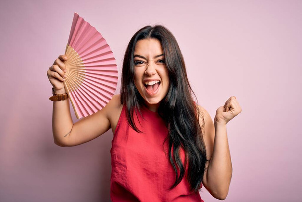 Young brunette woman holding hand fan for fresh air over pink isolated background screaming proud and celebrating victory and success very excited, cheering emotion - Photo, Image