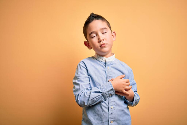 Young little boy kid wearing elegant shirt standing over yellow isolated background smiling with hands on chest with closed eyes and grateful gesture on face. Health concept. - Fotó, kép
