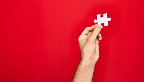 Beautiful hand of man holding piece of puzzle over isolated red background - Foto, afbeelding