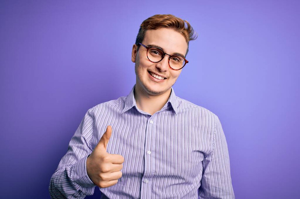 Young handsome redhead man wearing casual shirt and glasses over purple background doing happy thumbs up gesture with hand. Approving expression looking at the camera showing success. - Foto, Imagem
