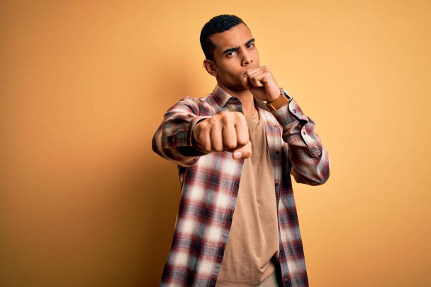 Молодий афроамериканець, одягнений в повсякденну сорочку, що стоїть над жовтим фоном Переказуючи кулак в бій, агресивний і сердитий напад, погроза і насильство. - Фото, зображення