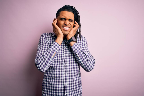 Ein junger gutaussehender afroamerikanischer Afro-Mann mit Dreadlocks trägt ein lässiges Hemd, das die Ohren mit Fingern bedeckt, die sich über den Lärm lauter Musik ärgern. Gehörloses Konzept. - Foto, Bild