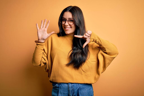 jonge brunette vrouw dragen bril en casual trui over geel geïsoleerde achtergrond tonen en wijzen met vingers nummer zeven terwijl glimlachen zelfverzekerd en gelukkig. - Foto, afbeelding