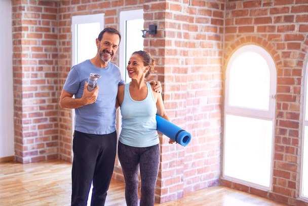 Middle age beautiful sporty couple smiling happy. Standing with smile on face hugging and holding mat at gym - Photo, Image