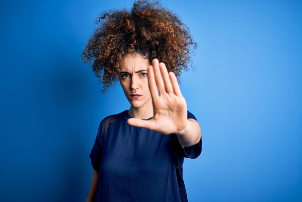 Eine junge schöne Frau mit lockigem Haar und Piercing trägt ein lässiges blaues T-Shirt und singt mit der Handfläche. Warnende Miene mit negativer und ernster Geste im Gesicht. - Foto, Bild