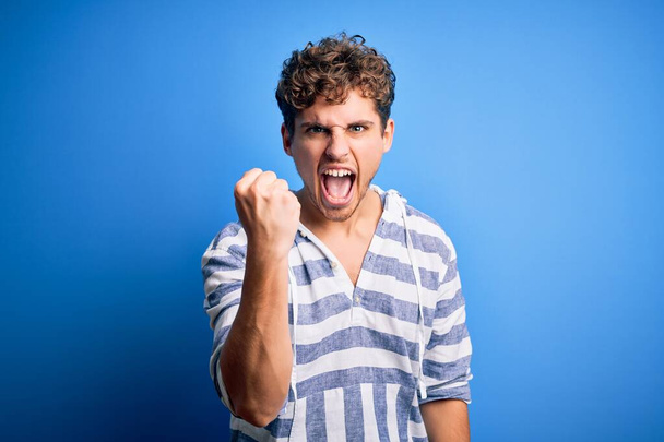 Young blond handsome man with curly hair wearing casual striped sweatshirt angry and mad raising fist frustrated and furious while shouting with anger. Rage and aggressive concept. - Foto, Imagen