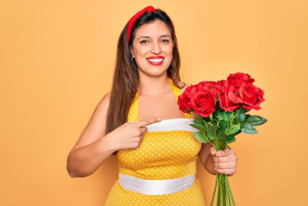 Jovem hispânico pin up mulher vestindo moda sexy estilo 50 segurando buquê de rosas vermelhas muito feliz apontando com mão e dedo
 - Foto, Imagem