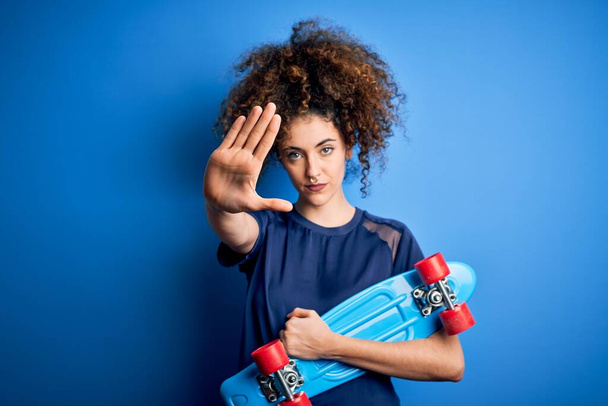 Young sporty woman with curly hair and piercing holding skate over blue background with open hand doing stop sign with serious and confident expression, defense gesture - 写真・画像