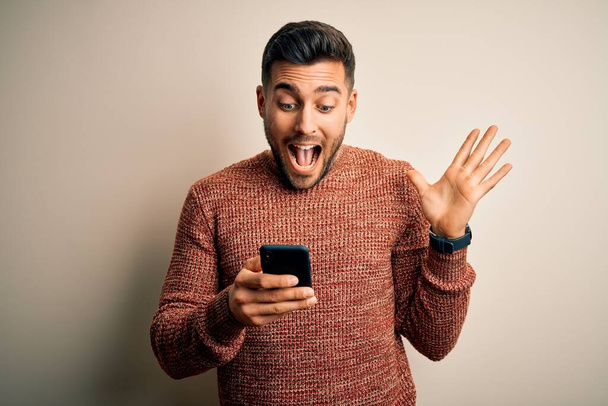 Young handsome man having conversation using smartphone over white background very happy and excited, winner expression celebrating victory screaming with big smile and raised hands - Fotó, kép
