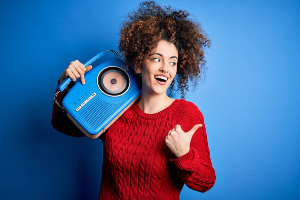 Jeune belle femme aux cheveux bouclés et perçant écoutant de la musique à l'aide d'une radio vintage pointant et montrant le pouce sur le côté avec un sourire heureux
 - Photo, image