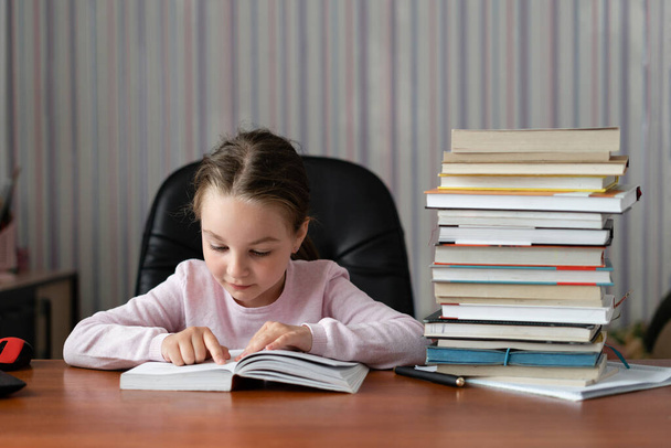 Ένα περίεργο κοριτσάκι, ένα παιδί με ευχαρίστηση, διαβάζει ένα βιβλίο και κάνει μαθήματα. Έννοια εκπαίδευσης σε σπίτι καραντίνας. - Φωτογραφία, εικόνα