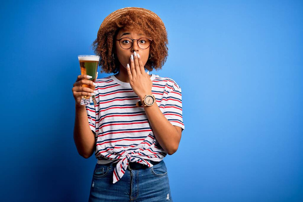 Jonge mooie Afro-Amerikaanse afro vrouw met krullend haar op vakantie drinken glas bier cover mond met de hand geschokt met schaamte voor fout, uitdrukking van angst, bang in stilte, geheim concept - Foto, afbeelding