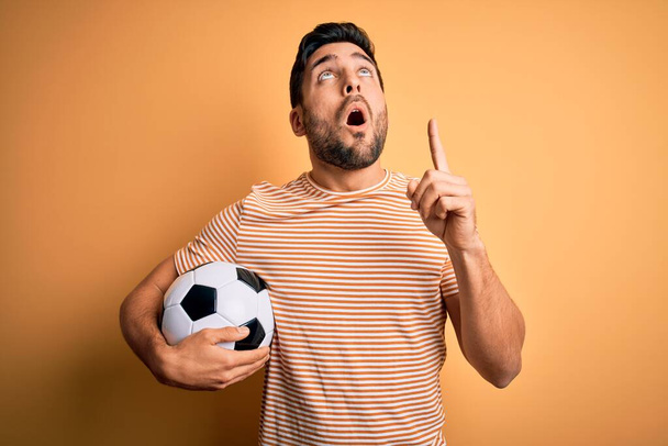 Jóképű, szakállas férfi focizik, focizik, focilabdát tart a sárga háttér felett, csodálkozva és meglepve néz fel, és ujjal mutogat, felemelt karral.. - Fotó, kép
