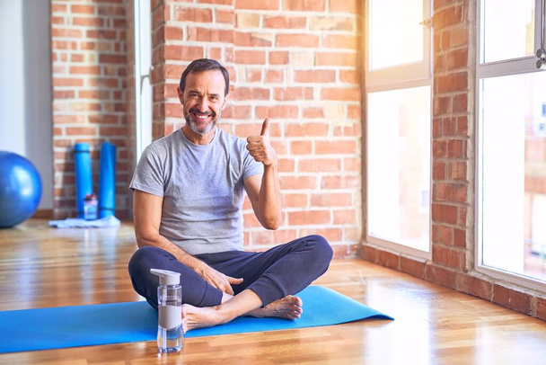 Przystojny sportowiec w średnim wieku siedzi na macie i ćwiczy jogę na siłowni robiąc szczęśliwy gest ręką w górę. Zatwierdzanie ekspresji patrząc w aparat pokazujący sukces. - Zdjęcie, obraz
