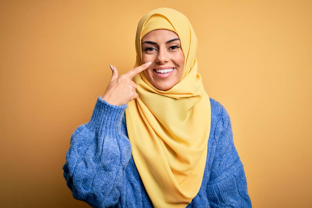 Fiatal gyönyörű barna muszlim nő visel arab hidzsáb felett elszigetelt sárga háttér mutató kéz ujját az arc és az orr, mosolygós vidám. Szépségkoncepció - Fotó, kép