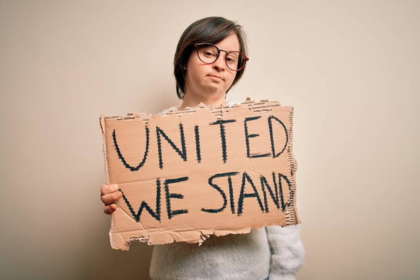 Young down syndrome woman holding protest banner with united we stand rights message with a confident expression on smart face thinking serious - Фото, изображение