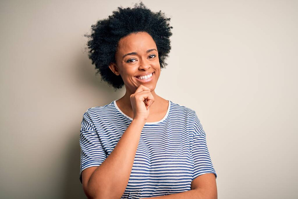 Jeune belle afro afro-américaine aux cheveux bouclés portant un t-shirt rayé regardant avec confiance la caméra avec le sourire aux bras croisés et la main levée sur le menton. Penser positif
. - Photo, image