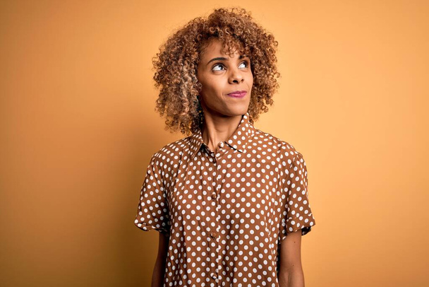 Junge schöne afrikanisch-amerikanische Frau in lässigem Hemd vor gelbem Hintergrund, lächelnd zur Seite schauend und denkend wegstarrend. - Foto, Bild